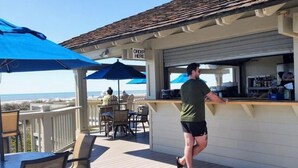 A bar area with a counter and a sign that says "ORDER HERE". There are outdoor tables and chairs, and a view of the ocean in the background.