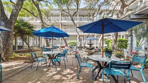 An outdoor patio with blue chairs and tables, surrounded by trees and a white building with balconies.