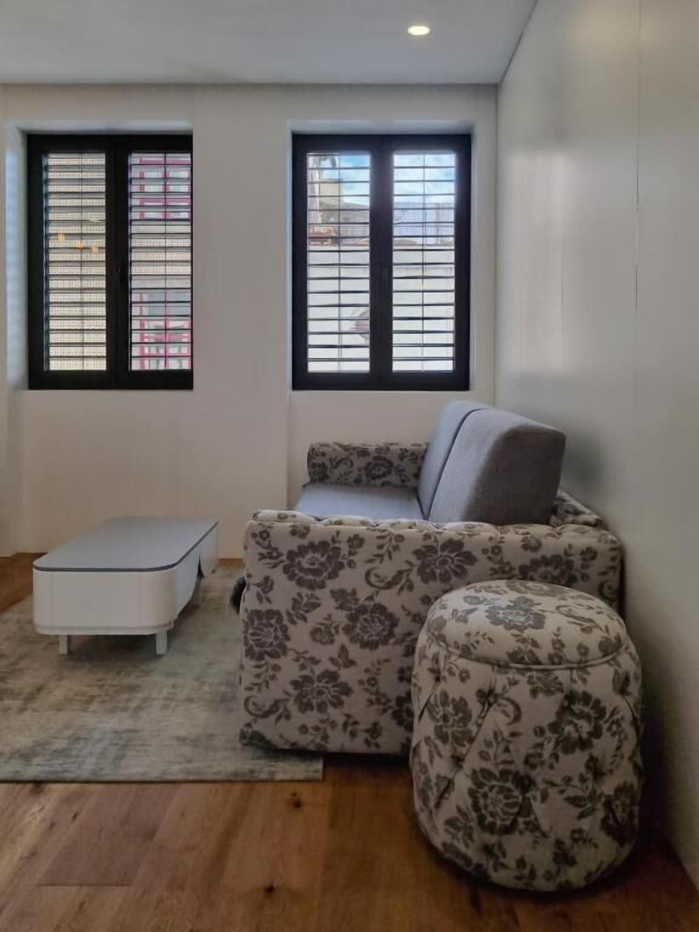 Une petite pièce agréable et chaleureuse, avec un canapé, un pouf et une petite table. Elle est dotée de deux fenêtres équipées de stores et le sol est recouvert de parquet.