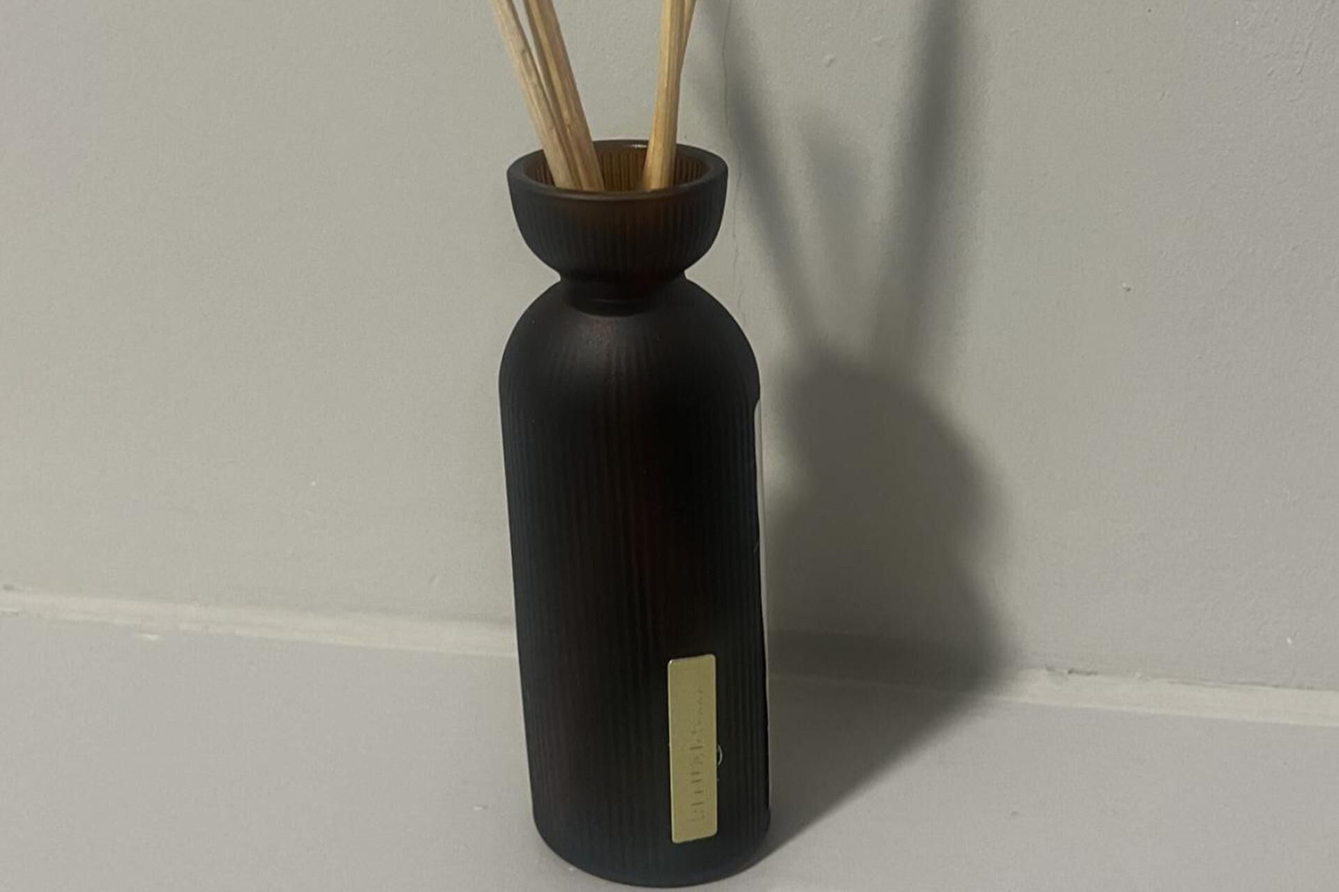 A black vase with a label on its side, placed on a flat surface against a plain wall.