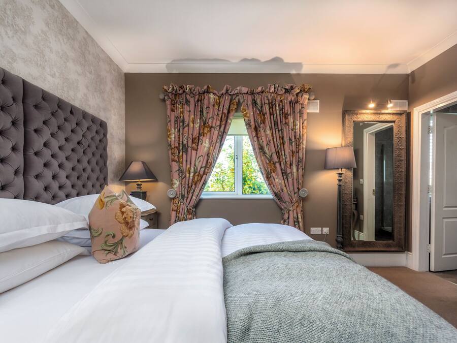 Un dormitorio con cabecera capitonada, una cama grande con ropa de cama blanca, un plaid estampado floral y una ventana con cortinas florales.