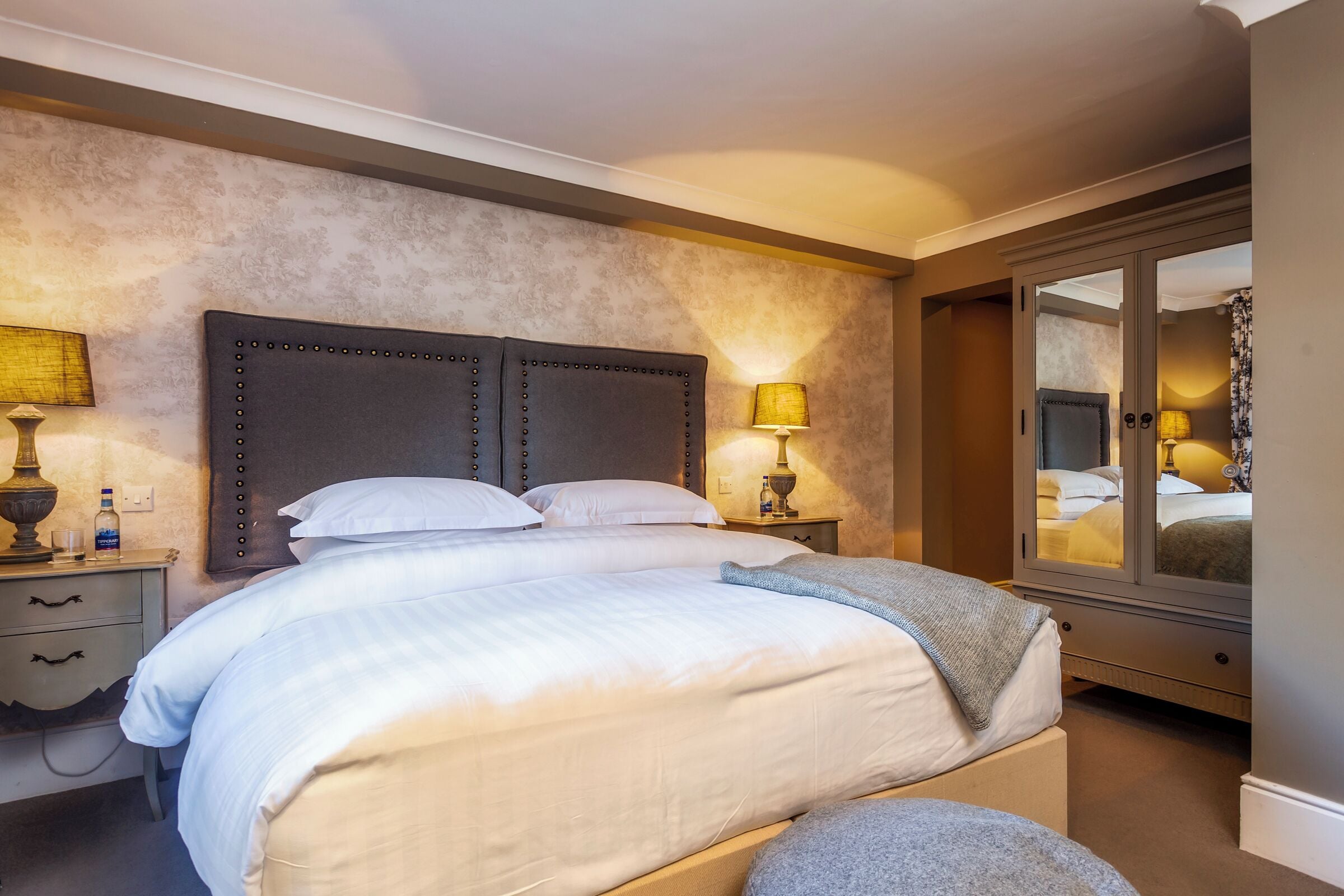 Una cama bien hecha con cabecera, lámparas de noche, mesitas y vistas a otra habitación a través de un ventanal.
