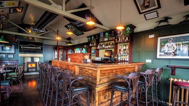 Una zona de bar con taburetes de madera, un mostrador con una caja registradora y una variedad de botellas de licores.