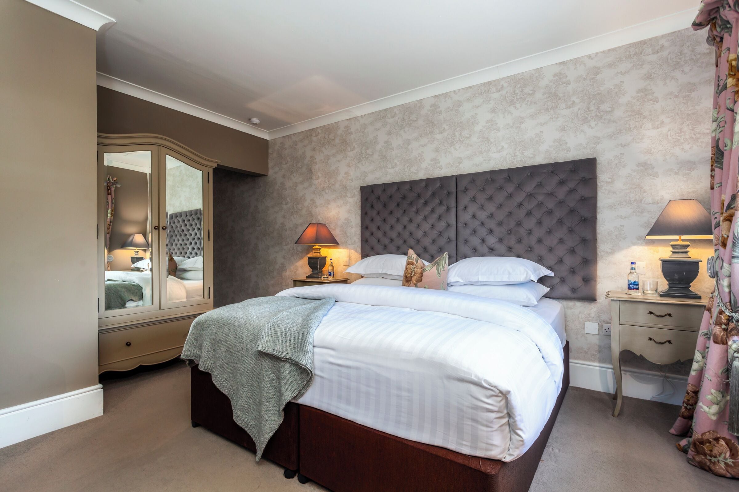 A bedroom with a large bed, a wardrobe, a bedside table with a lamp, and a mirror.