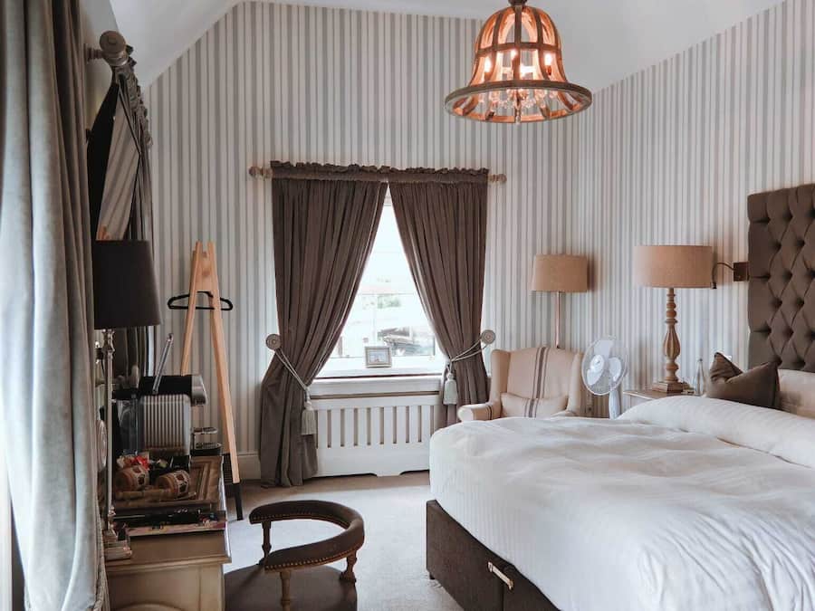Habitación de hotel con una cama grande, una zona de estar con una silla, un escritorio con lámpara y un ventanal con cortinas.
