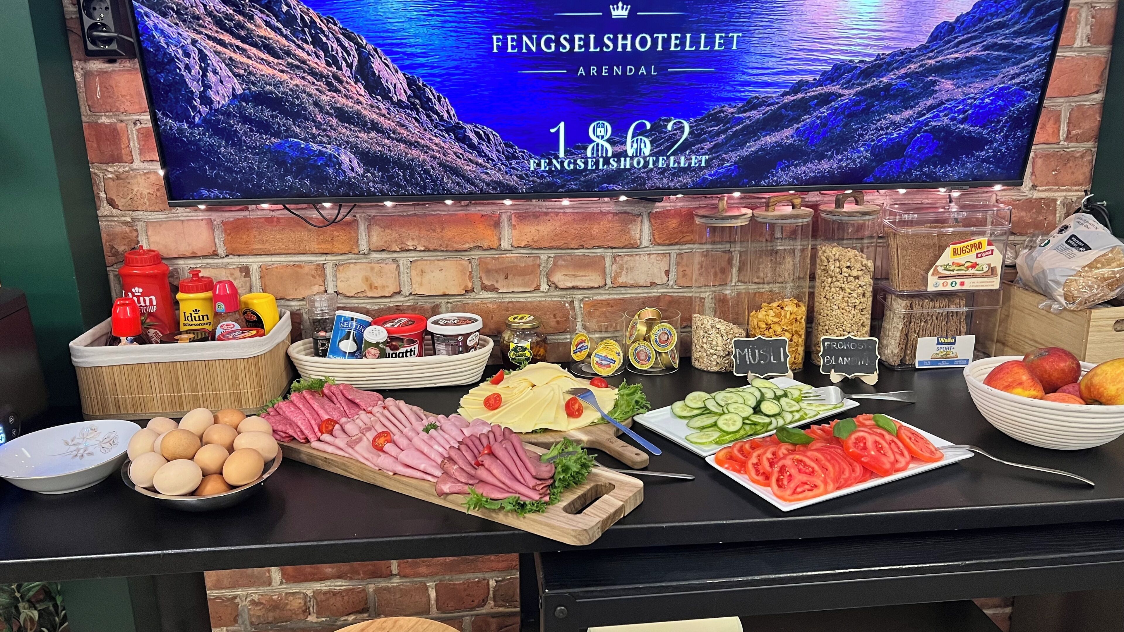 Mesa de buffet con diversas opciones de comida, salsas y un televisor mostrando el nombre y año de fundación del hotel.