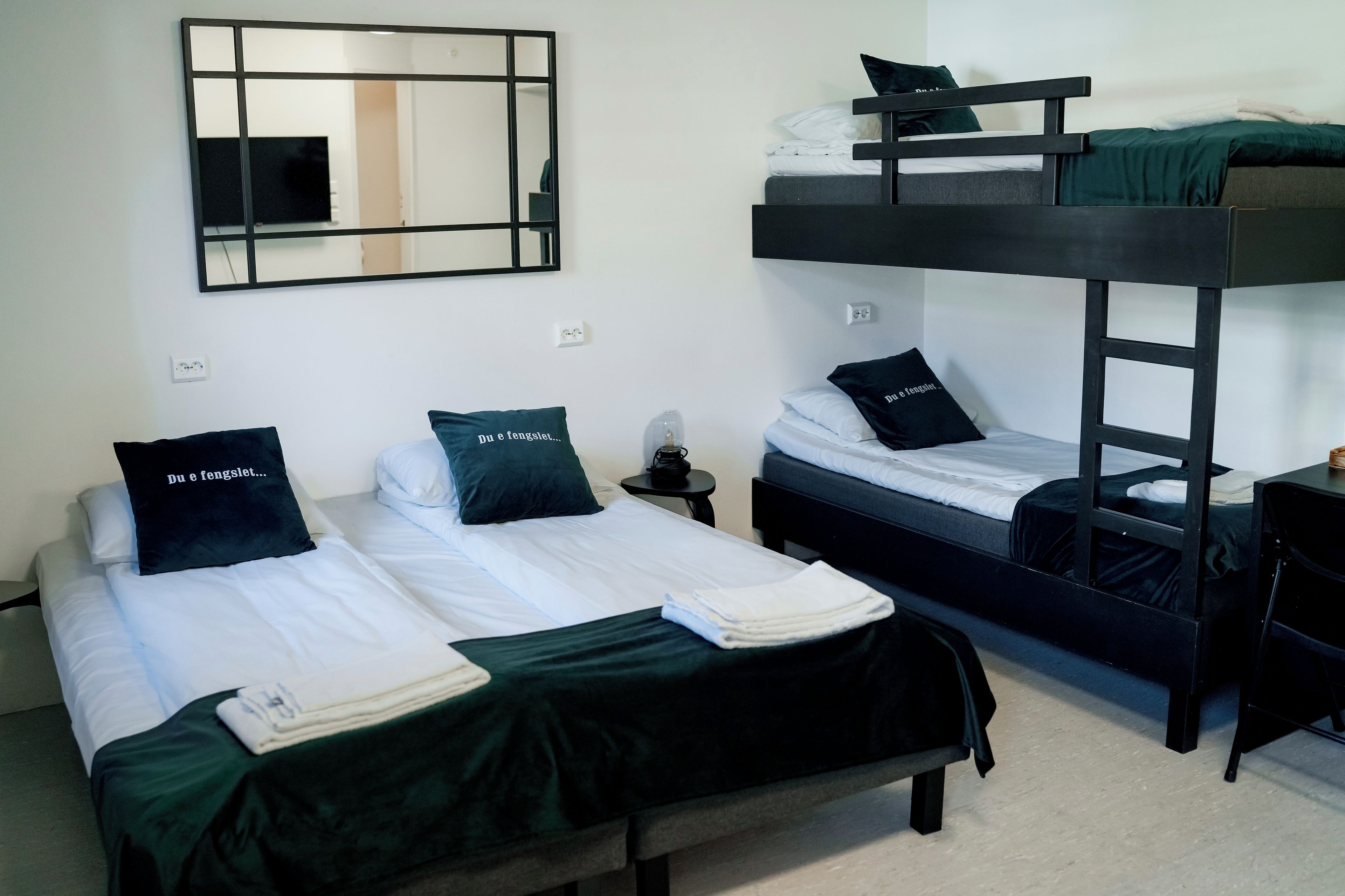 A room with bunk beds, a desk, a chair, a mirror, and a bedside table with a lamp.
