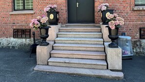 A brick building with a black double door, steps leading up to the entrance, and two flower pots with pink flowers on either side. - Fengselshotellet (Arendal)