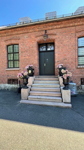 A brick building with a black double door, steps leading up to the entrance, and two flower pots with pink flowers on either side. - Fengselshotellet (Arendal)