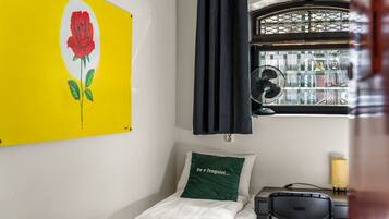 Een kleine kamer met een bed, een stoel, een bureau, een raam met uitzicht op gebouwen, en een wand met een roos schilderij.