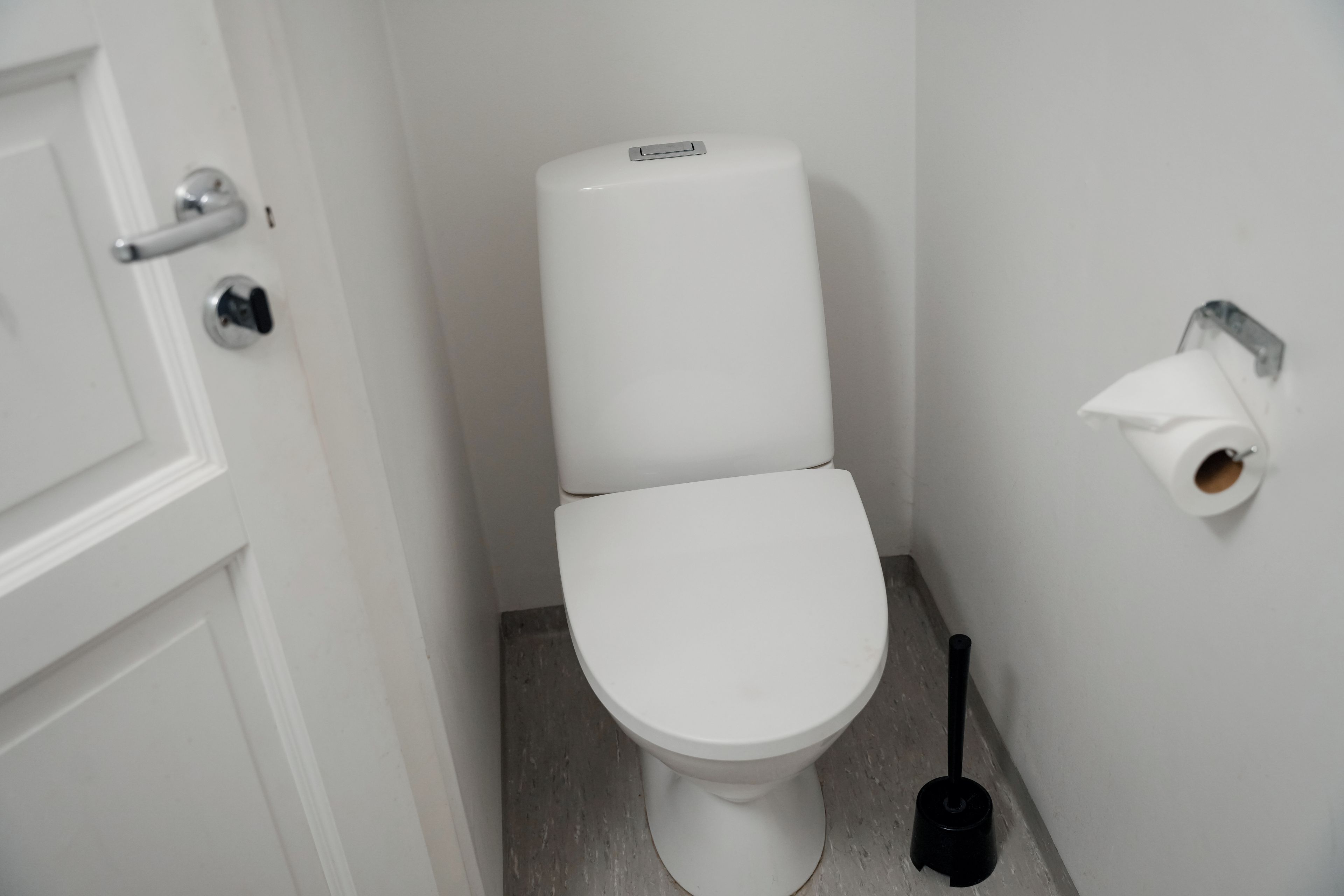 A white toilet with a flush handle, a toilet paper holder, and a plunger on the floor.