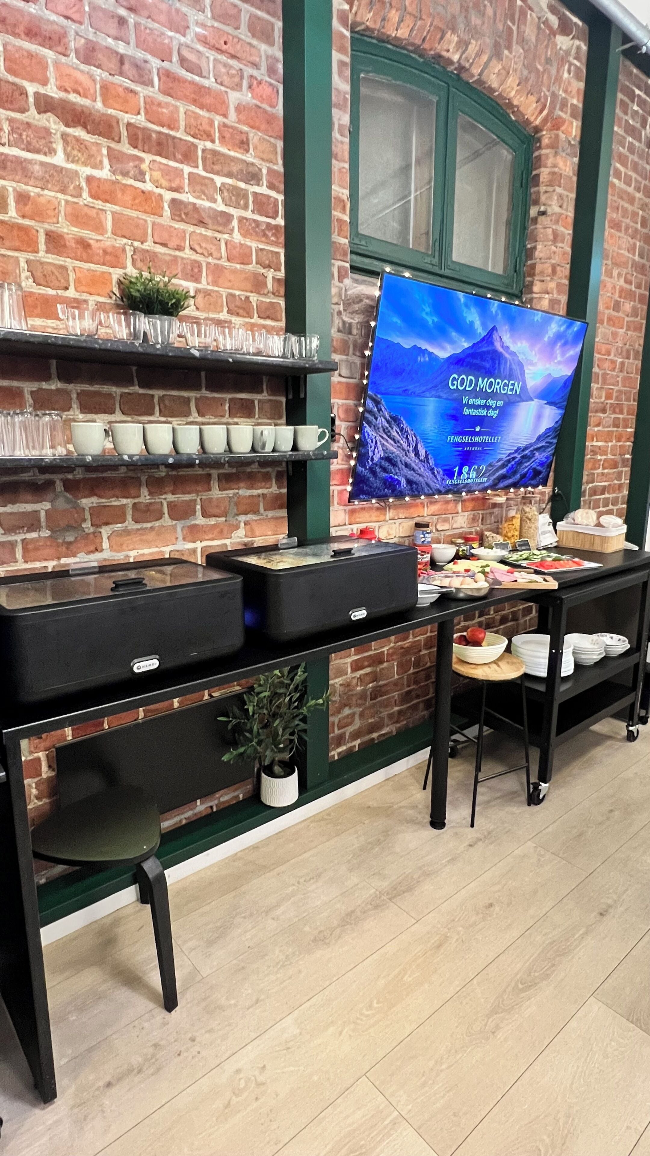 Pared de ladrillo con una ventana de marco verde, un televisor de pantalla plana mostrando un paisaje montañoso y una mesa de buffet con diversos alimentos y utensilios.