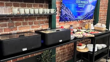 Pared de ladrillo con una ventana de marco verde, un televisor de pantalla plana mostrando un paisaje montañoso y una mesa de buffet con diversos alimentos y utensilios.