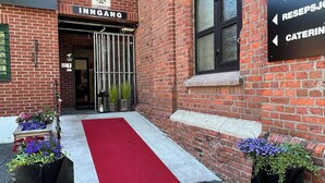 A brick building with a red carpet entrance, a sign for 'INNGANG,' and directional signs for 'SELSKAP LOKALER,' 'RESEPSJOG,' and 'CATERING.' - Fengselshotellet (Arendal)