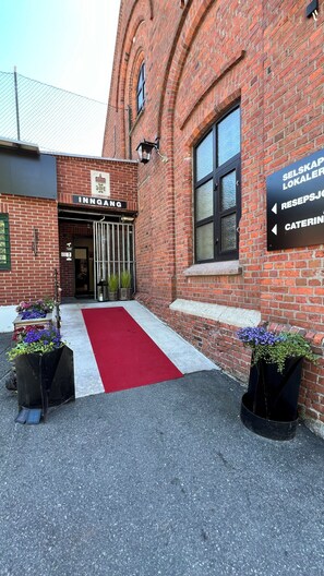 Edificio de ladrillo con alfombra roja en la entrada, un cartel que dice 'INNGANG' y señales que indican 'SELSKAP LOKALER', 'RESEPSJOG' y 'CATERING'.