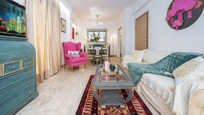 A living room with a teal dresser, a pink armchair, a white sofa, and a glass coffee table.
