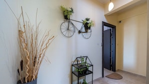 A hallway with a wall-mounted bicycle planter, a small shelf with potted plants, and a tall vase with reeds.