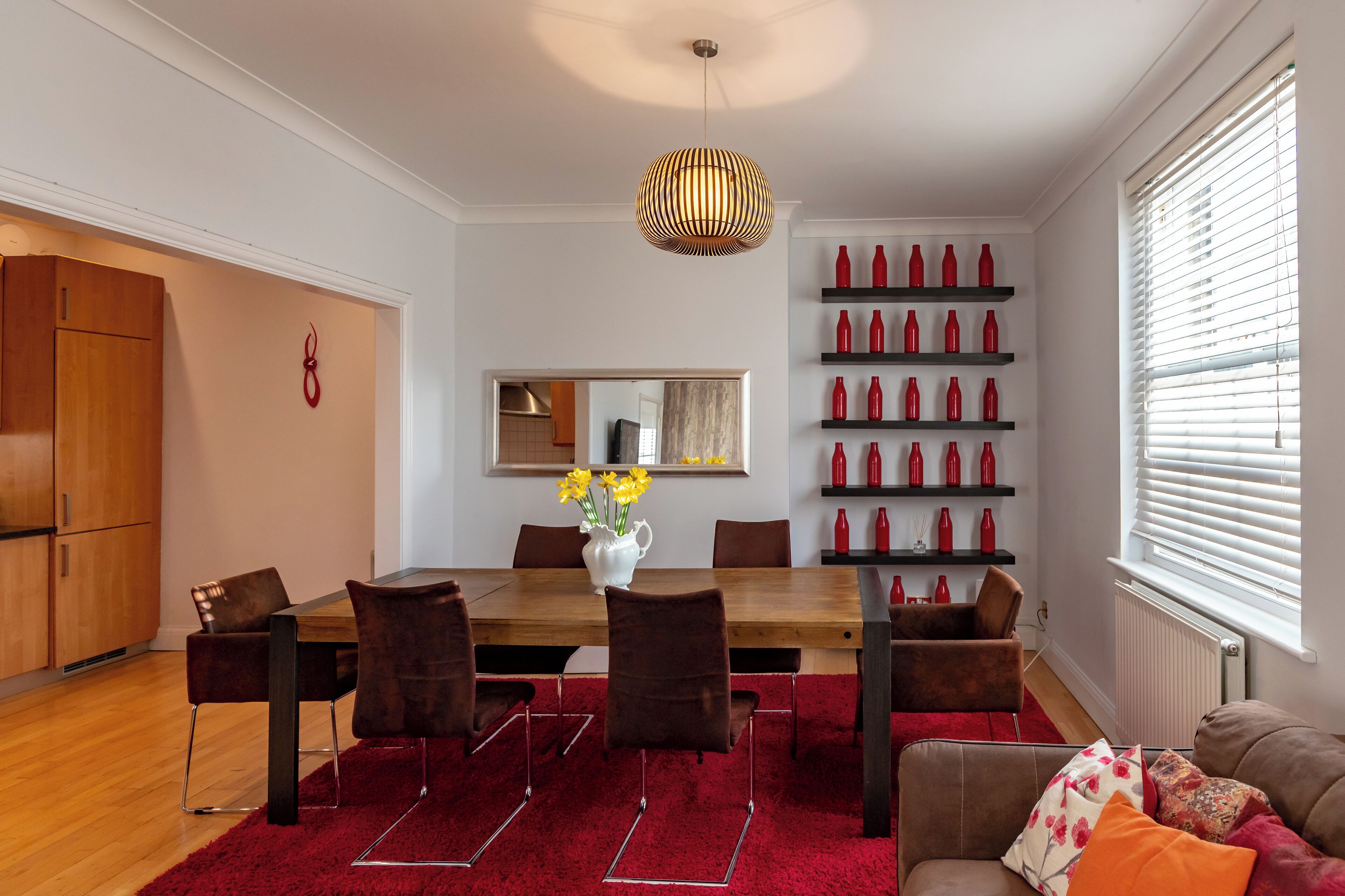 Uma sala de jantar moderna com mesa de madeira, cadeiras marrons e tapete vermelho. Há um vaso com flores amarelas sobre a mesa.