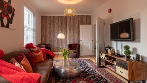 Ein modernes Wohnzimmer mit einem braunen Sofa, roten Kissen, einem runden Couchtisch mit einer Vase, einem gemusterten Teppich, einer Holzkommode und einem an der Wand befestigten Fernseher.