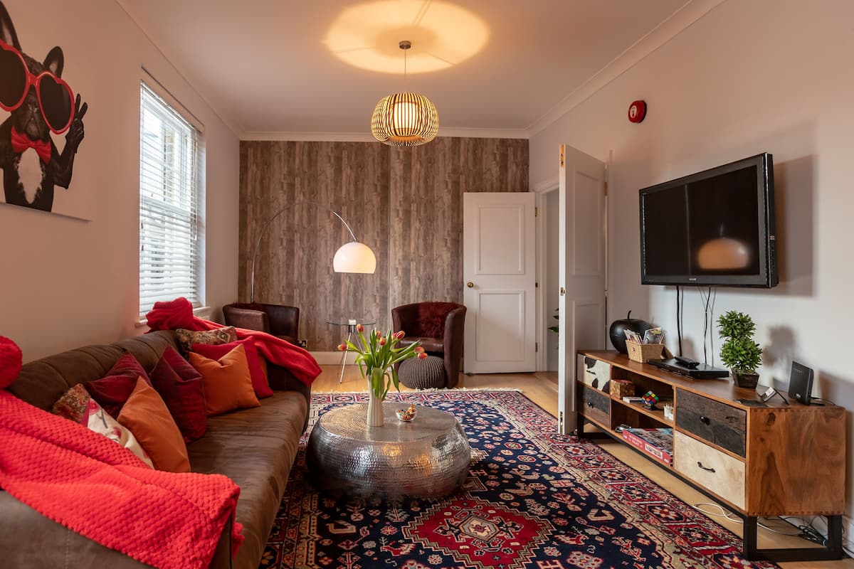 A modern living room with a brown sofa, red cushions, a round coffee table with a vase, a patterned rug, a wooden cabinet, and a mounted TV.