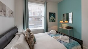 Ein modernes Hotelzimmer mit einem großen Bett, einem Nachttisch mit Lampe, einem Fenster mit Vorhängen und Kunstwerken an den Wänden.