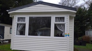 A small, single-story mobile home with a gabled roof and two windows with curtains. - Immaculate 3-bed Caravan 10mins From Gt Yarmouth (Great Yarmouth)