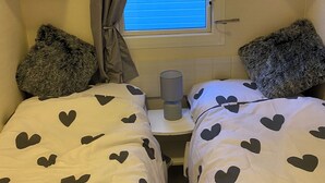 A small, cozy room with two beds, each with a white duvet featuring black heart patterns. There is a window with a view of a building, and a small table with a lamp and a roll of toilet paper. - Immaculate 3-bed Caravan 10mins From Gt Yarmouth (Great Yarmouth)