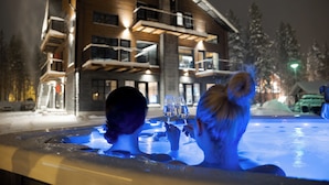Een ruimte met een hottub, twee personen, een houten gebouw met balkons en een besneeuwde omgeving.