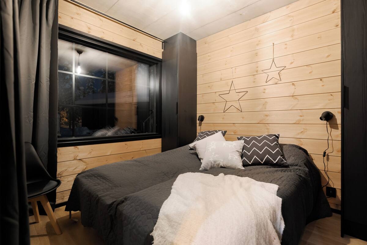 Une chambre avec une grande fenêtre, des murs en bois, un lit avec de nombreux oreillers et un fauteuil.