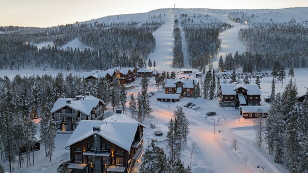 Een met sneeuw bedekte lodge met meerdere gebouwen, een piste op de achtergrond en een bebost gebied.