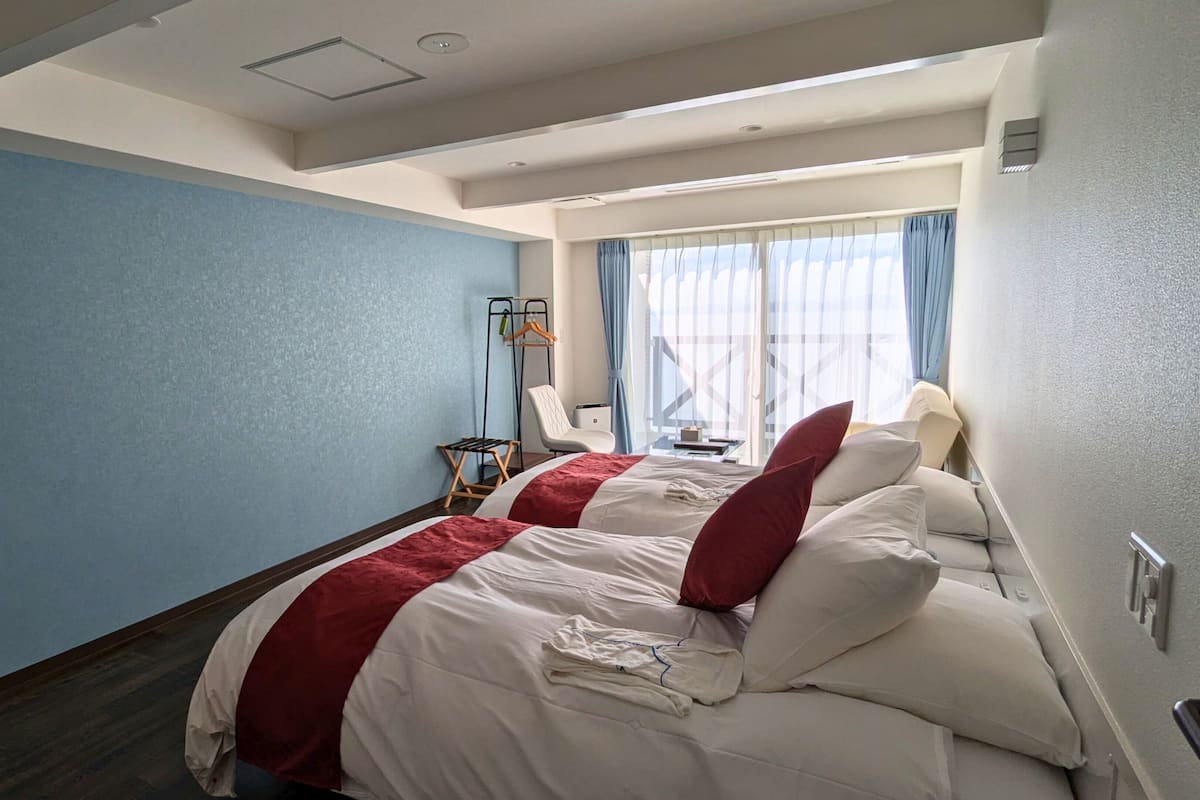 Une chambre d’hôtel avec deux lits, une chaise, une petite table et une fenêtre avec des rideaux.