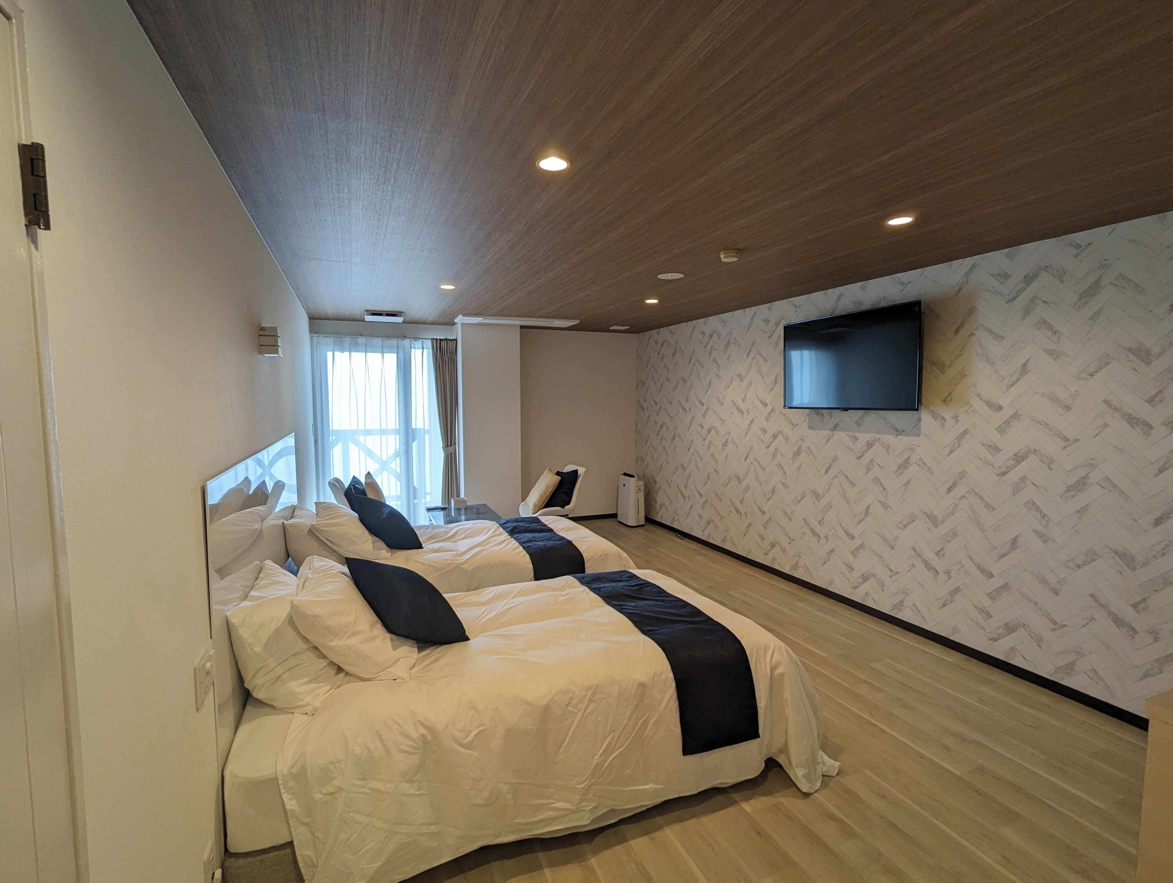 Ein modernes Hotelzimmer mit zwei Betten, einem an der Wand befestigten Flachbildfernseher und einem großen Fenster für natürliches Licht.