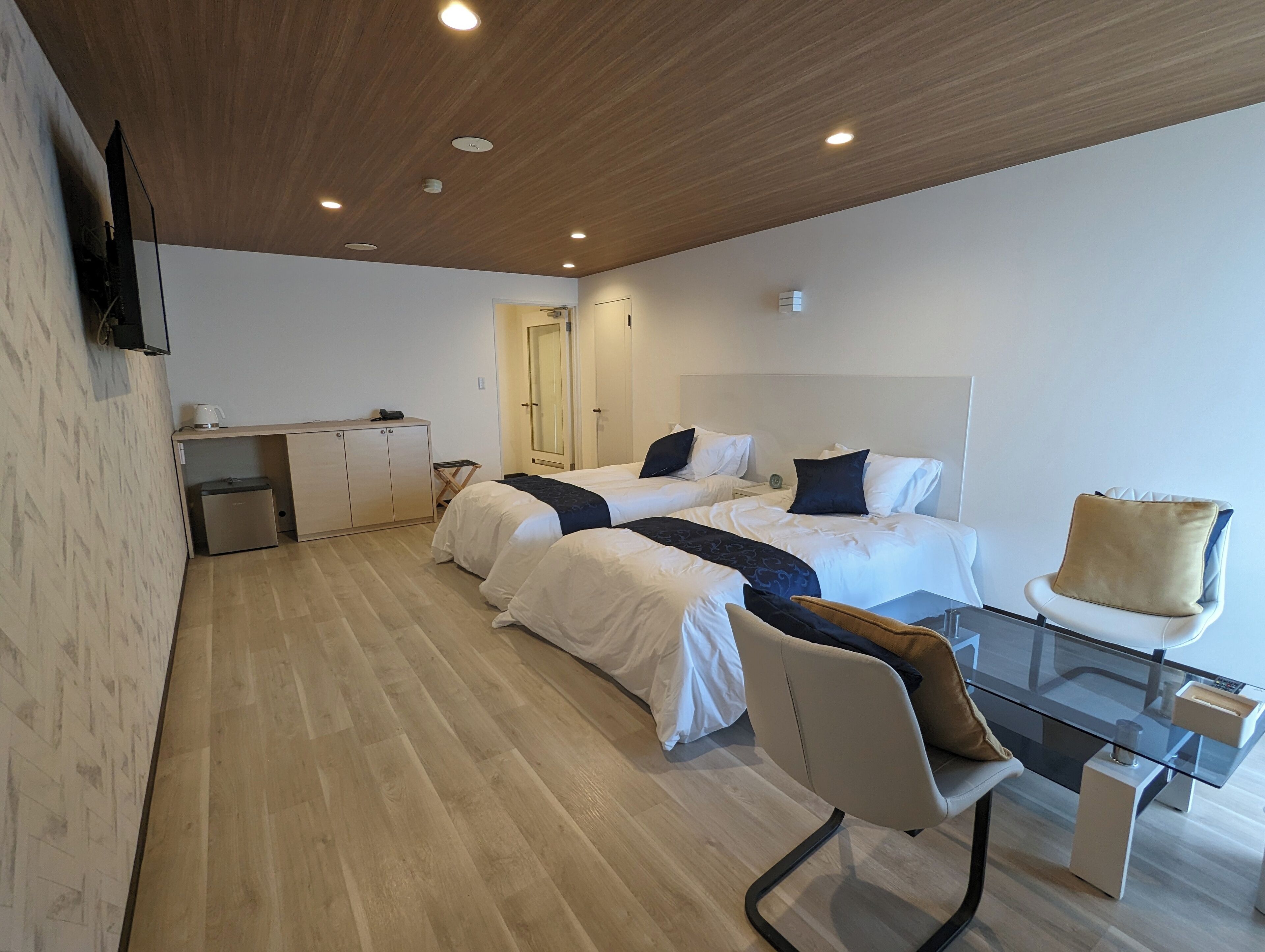 A modern hotel room with two beds, a small desk, a glass table, and a wooden ceiling.