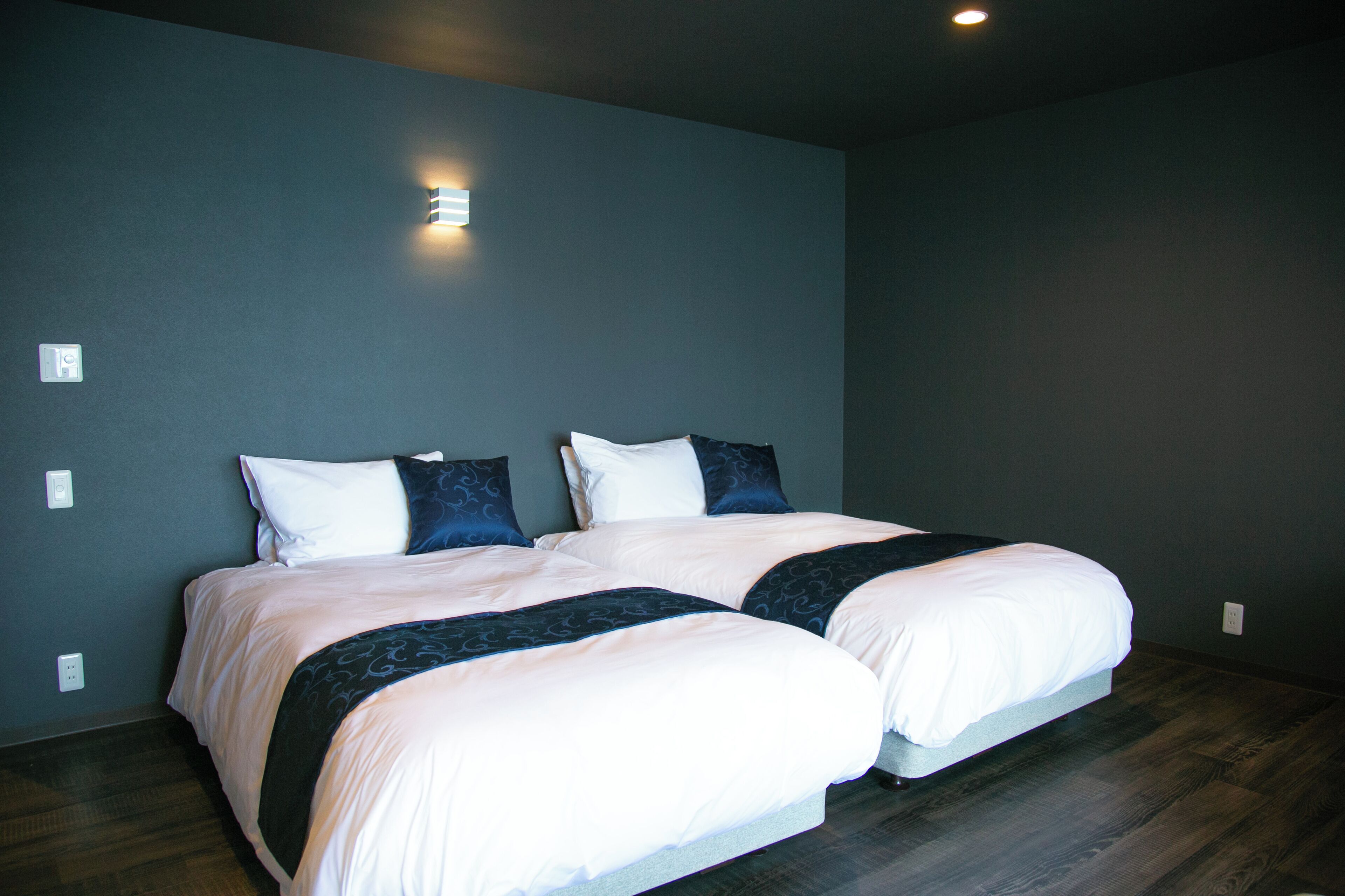 A double bed with white and dark blue bedding, a wall-mounted light, and a dark wooden floor.