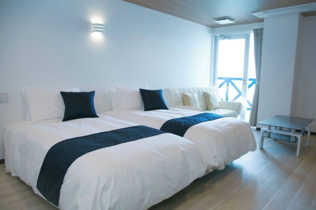 A hotel room with two beds, a small table, and a balcony with a railing.