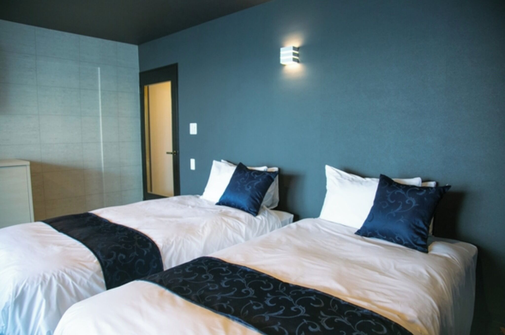 A hotel room with two beds, a door, and a wall-mounted light fixture.