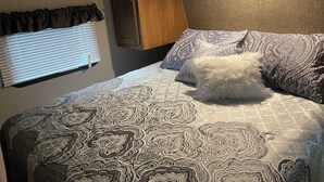 A bed with patterned bedding, a window with blinds, and a wall-mounted fire alarm. - Nature's Haven Tiny Home (Bear)