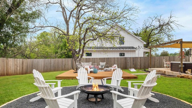Ein Garten mit Feuerstelle, weißen Adirondack-Stühlen, einer Holzterrasse und einem weißen Haus im Hintergrund.