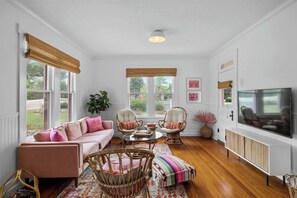 Una habitación con un sofá, dos sillas de mimbre, una mesa de centro, un televisor y ventanas grandes.