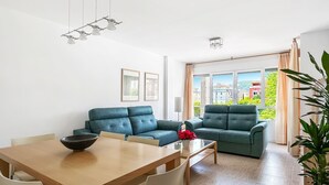 A modern living room with a sofa, a dining table, and a large window with a view of buildings and greenery. - Apartamento Solamaza con Terraza XL (Arnuero)