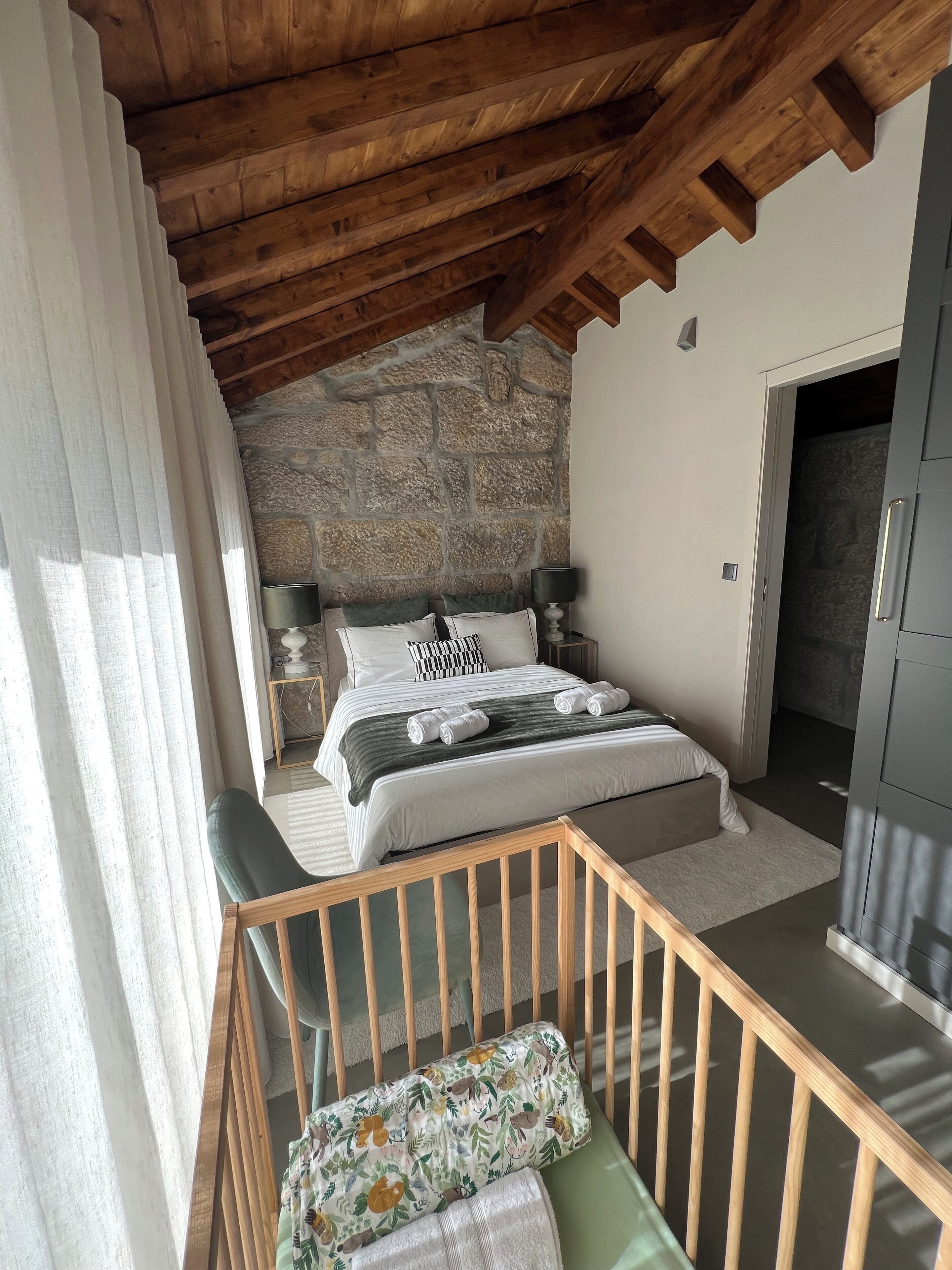 A bedroom with a stone wall, wooden ceiling beams, a bed with pillows, a bedside table, and a green chair.
