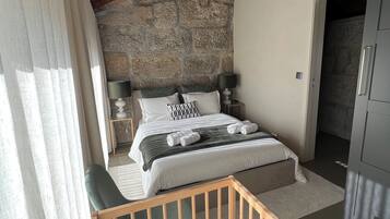 A bedroom with a stone wall, wooden ceiling beams, a bed with pillows, a bedside table, and a green chair.