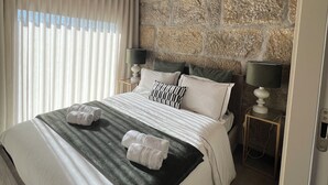 A bedroom with a stone wall, wooden ceiling beams, a bed with white linens, two bedside lamps, and a window with white curtains.