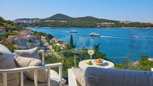 A balcony with a glass barrier overlooking a coastal town, a glass of wine, and a plate with food. - Hedera Estate, Hedera A25 (Dubrovnik)