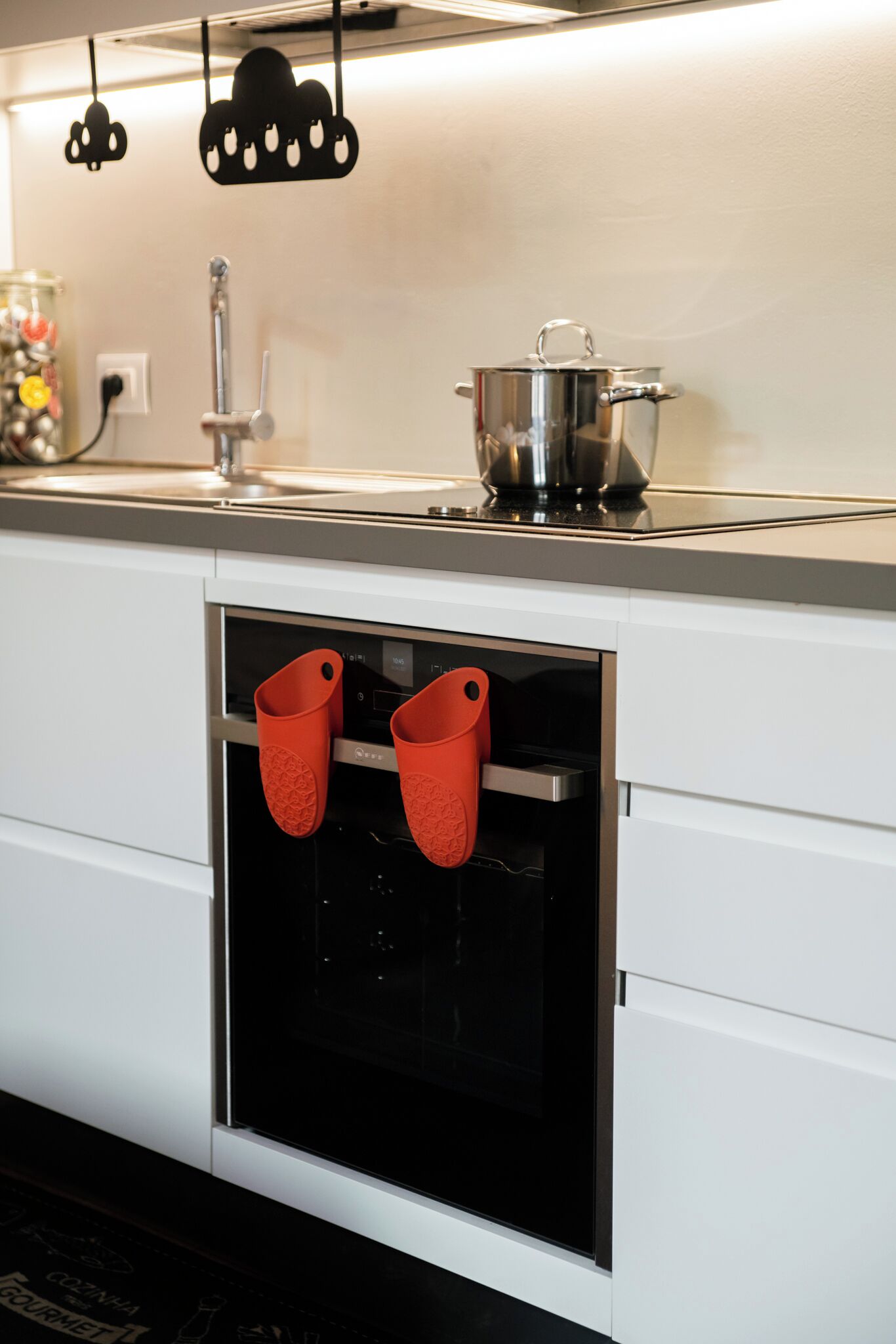 A kitchen with a stainless steel pot on the counter, a black oven door, and two red oven mitts hanging from the oven handle.