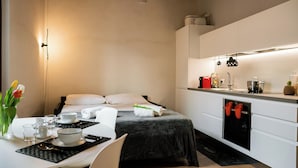 Une chambre d’hôtel compacte comprenant un lit, une table de chevet avec un vase de fleurs, une petite table à manger avec un bol et une cuillère, et une kitchenette équipée d’un réfrigérateur et d’une machine à café.