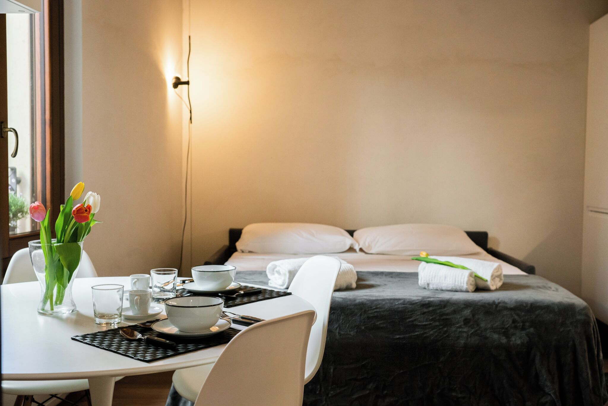 A hotel room with a neatly made bed, a dining table with a vase of tulips, and a window with a view.