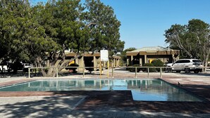 Una piscina con borde de ladrillo, rodeada de árboles y un edificio con un letrero.