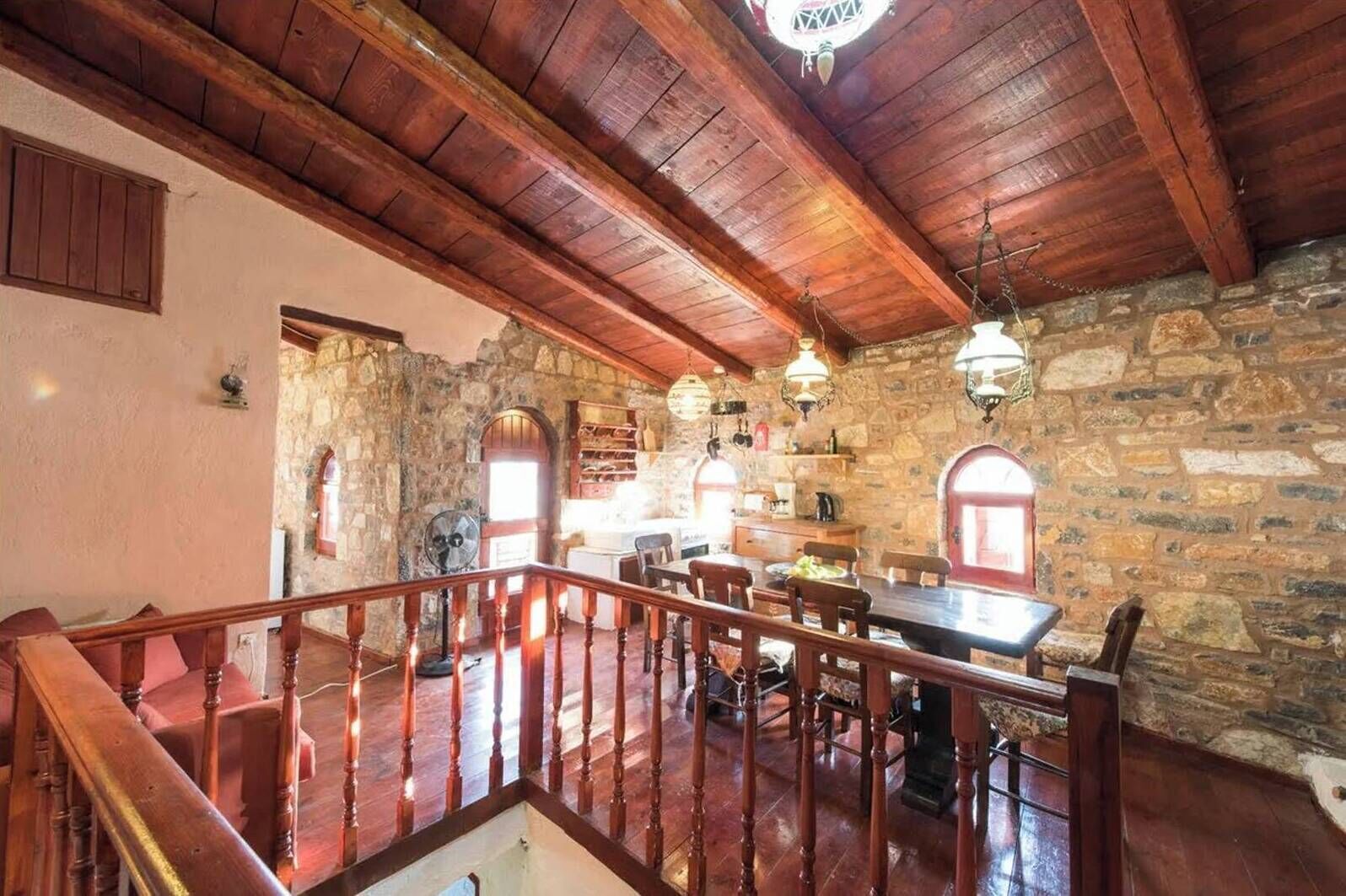 A rustic interior with a stone wall, wooden ceiling beams, a dining area with chairs and a table, and a staircase with wooden railings.