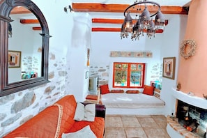 A cozy room with a stone wall, a wooden ceiling, a fireplace, a sofa, a bed, and a large window with a view. - Elounda Collection Villa (Agios Nikolaos)